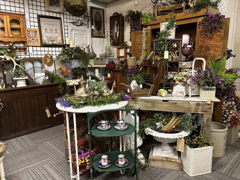 Garden display with potted flowers, a white plant stand, teacups on a green tiered stand and a wooden hutch
