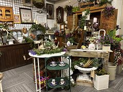 Garden display with potted flowers, a white plant stand, teacups on a green tiered stand and a wooden hutch