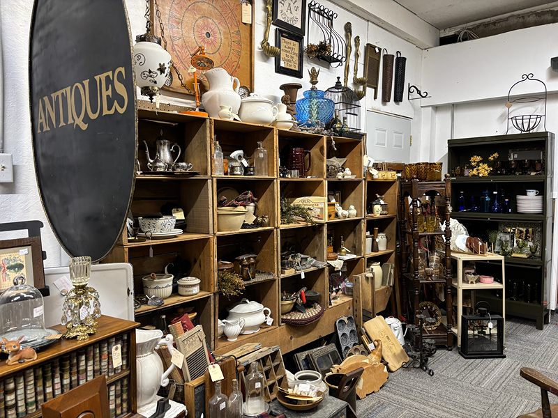 An Antiques sign beside wooden cubbies full of white pitchers, silver and kitchenware