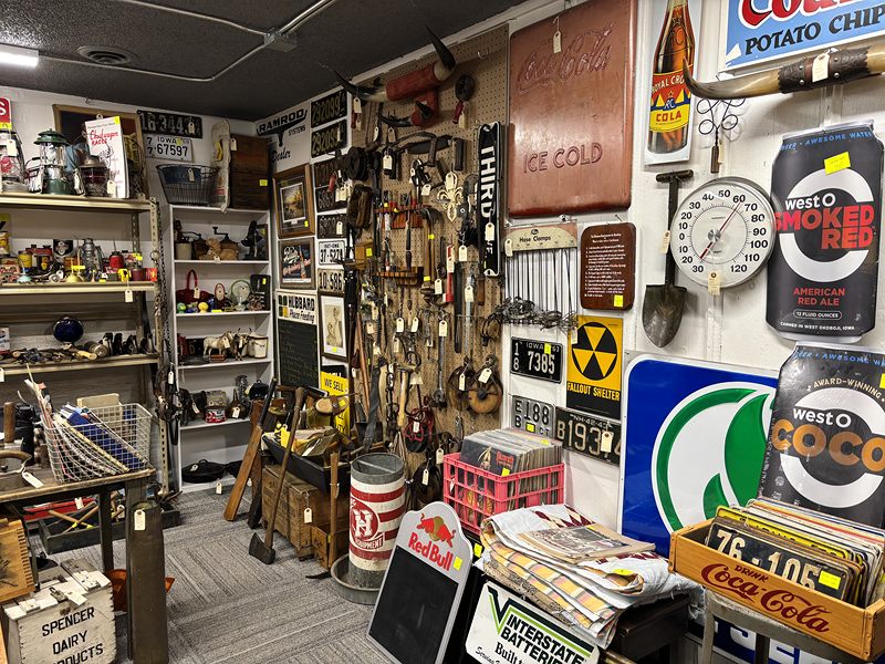 Wall of old tools and advertising signs, including Coca-Cola, above shelves of collectibles