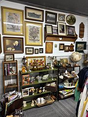 Wall of framed prints and paintings above shelves of glassware and knick-knacks