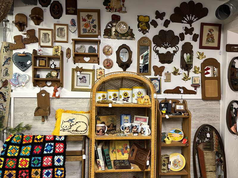 Wall of wooden decor and framed needlework above a wicker shelf and a granny-square afghan