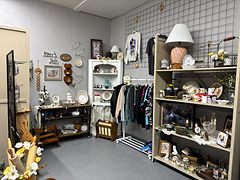 Booth with a white hutch of dishes, a rack of clothes, a lamp and shelves of home decor