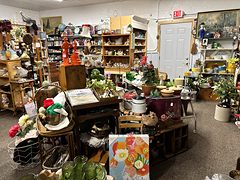 Busy aisle of antiques with orange glass, potted plants, baskets and a floral painting