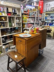 Wooden sewing cabinet and stool in an aisle lined with shelves of dishes and decor